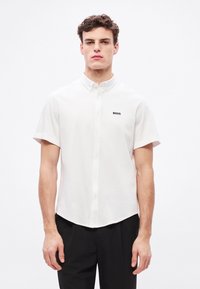 Balti trumpomis rankovėmis medvilniniai marškiniai, su sagutėmis užsegamu apykakliu ir mažu juodu „BOSS“ logotipu kairėje krūtinės pusėje. Prigludęs fasonas.