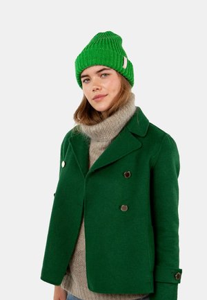 Green wool double-breasted coat with silver buttons, layered over a grey knitted turtleneck and a bright green ribbed beanie.