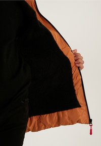 Interior de chaqueta negra con forro naranja, que presenta un tejido suave y texturizado. Cierre frontal con cremallera y un detalle en color rojo.