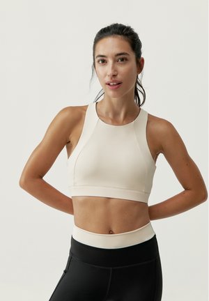 Sujetador deportivo de beige claro con un diseño ajustado, tirantes anchos y una textura suave, combinado con leggings negros de tiro alto.