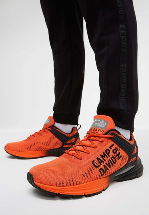 Orange running shoes with black accents, mesh upper, textured design, bold branding, and cushioned soles visible with black joggers.