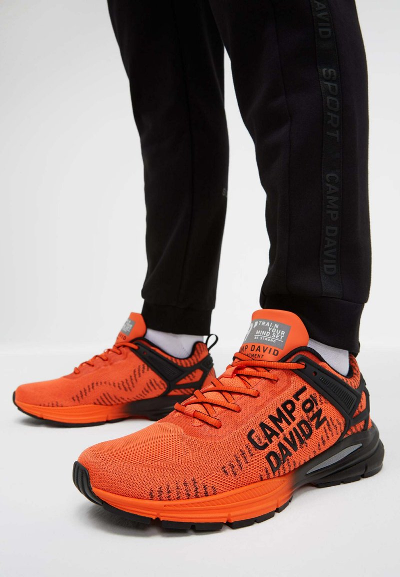 Orange Laufschuhe mit schwarzen Akzenten, Obermaterial aus Mesh, strukturiertes Design, markante Markenkennung und gepolsterte Sohle, sichtbar mit schwarzen Jogginghosen.