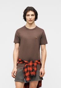 Νέος άντρας με μεσαίου μήκους σκούρα μαλλιά, που φοράει ένα καφέ t-shirt και ένα κόκκινο-μαύρο καρό πουκάμισο δεμένο γύρω από γκρι σορτς.