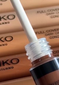 Anti-cernes à couvrance totale dans un tube transparent avec une bande noire, muni d'un applicateur blanc. En arrière-plan, un emballage de produit beige.