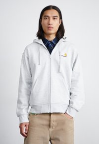 Sweat à zip gris avec cordons, arborant un petit logo brodé sur la poitrine. Porter par-dessus une chemise à carreaux bleue, avec des poches latérales.
