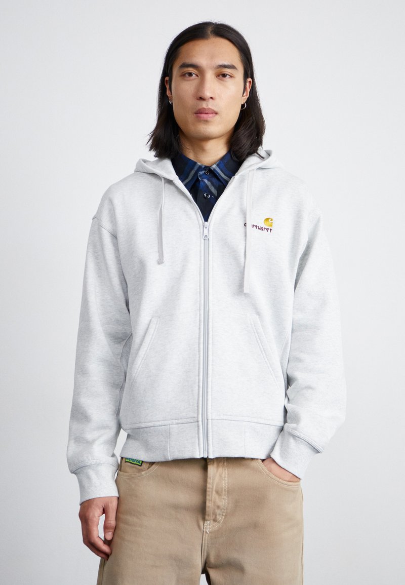Sweat à zip gris avec cordons, arborant un petit logo brodé sur la poitrine. Porter par-dessus une chemise à carreaux bleue, avec des poches latérales.
