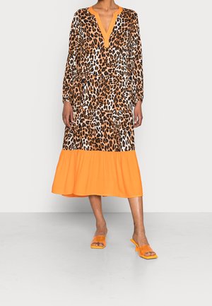 Nainen, joka pukeutunut leopardikuvioiseen midimekkoon, jossa oranssi helma ja kaula-aukko, yhdistettynä oransseihin avokärkisiin korkokenkiin.