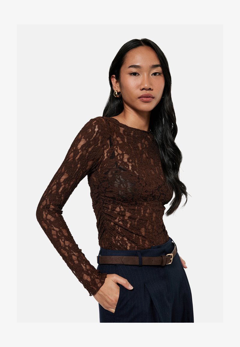 Femme aux longs cheveux noirs portant un haut ajusté en dentelle marron à manches longues, un pantalon foncé avec une ceinture marron et des boucles d'oreilles anneaux dorées.