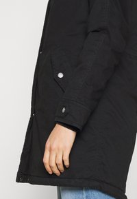 Svart parka-jacka med texturerat tyg, tryckknappdetaljer och sidofickor, visad från sidan med en delvis synlig hand.