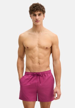 Man zonder shirt met krullend haar en felmagenta zwemshorts, staand met handen in de zakken tegen een lichte, effen achtergrond.