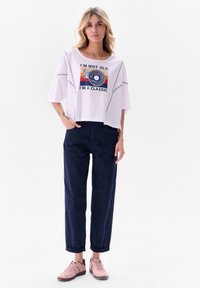 T-shirt blanc court avec un graphisme coloré indiquant "JE NE SUIS PAS VIEUX, JE SUIS UN CLASSIQUE", accompagné d'un pantalon bleu marine et de baskets roses.