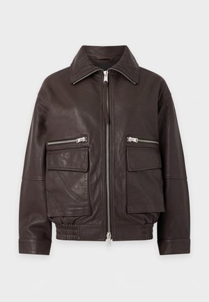 Un mannequin se tient devant un fond uni, vêtu d’une veste en cuir marron foncé, d’un pantalon noir évasé, de bottes pointues, de créoles et d’un collier à pendentif.