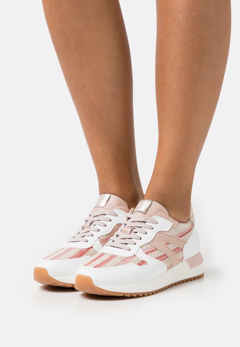 Personne portant des baskets rose clair et blanches avec un motif texturé et des semelles en gomme, debout sur un fond clair uni.