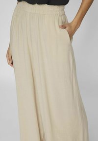 Pantalones de pierna ancha color beige claro con cintura elástica, que cuentan con bolsillos laterales y una textura de tejido suave y fluido.