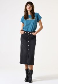 T-shirt teal avec le mot "MORE" en lettres dorées, associé à une jupe midi en denim noir avec des boutons sur le devant et des poches, portée avec des bottines noires.
