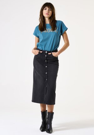 T-shirt teal avec le mot "MORE" en lettres dorées, associé à une jupe midi en denim noir avec des boutons sur le devant et des poches, portée avec des bottines noires.