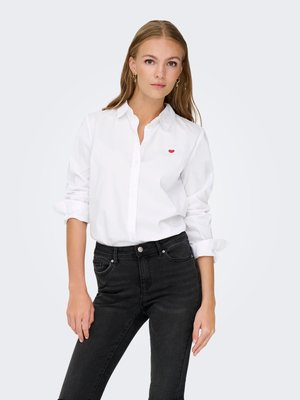 Giovane donna con capelli lunghi che indossa una camicia bianca abbottonata con un dettaglio a cuore rosso e jeans neri, in piedi davanti a uno sfondo chiaro e semplice.
