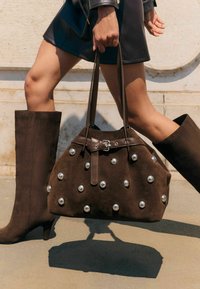 Barna nubuk bőr kézitáska ezüst szegecsekkel, lekerekített forma, dupla fogantyúkkal és csattos részlettel. Térd fölé érő barna nubuk bőr csizmával viselve.