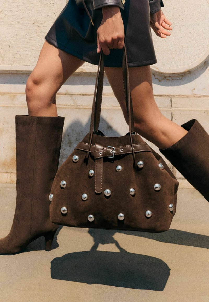 Barna nubuk bőr kézitáska ezüst szegecsekkel, lekerekített forma, dupla fogantyúkkal és csattos részlettel. Térd fölé érő barna nubuk bőr csizmával viselve.