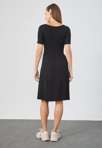 Schwarzes knielanges Kleid mit kurzen Ärmeln und tailliertem Oberteil, das eine geraffte Taille und einen weichen Stoff aufweist; kombiniert mit hellen Sneakers.