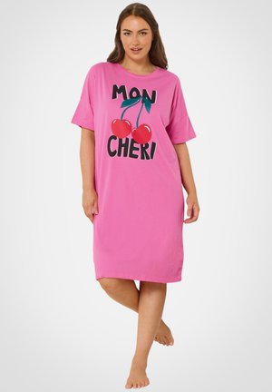 Kvinde stående barfodet iført en løs rosa kjole med to røde kirsebær og teksten "MON CHERI" på forsiden.