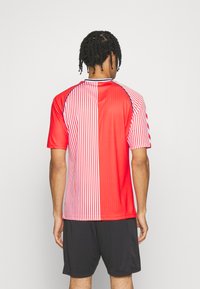 Punapääinen mies, jolla on letitetyt hiukset, seisoo selin päin käyttäen punavalkoraidallista pystyraitaista urheilupaitaa ja mustia shortseja tasaisella taustalla.