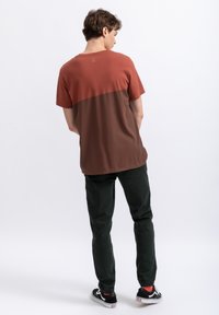 Heren T-shirt in een roest- en bruinkleurig blokontwerp, korte mouwen, ronde halslijn en logodetail op de bovenrug. Gekleed met donkere groene broek.