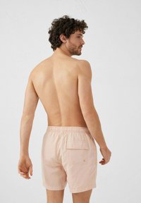 Pantaloni da bagno rosa chiaro con vita elastica e tasca posteriore, indossati da un uomo in piedi con le spalle rivolte verso l'osservatore.