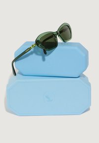 Zielone, oversized okulary przeciwsłoneczne w kształcie kociego oka, wyeksponowane na dwóch jasnoniebieskich etui. Ramiona mają dekoracyjne zielone ozdoby.