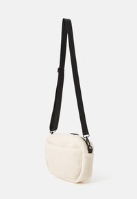 Krémová crossbody taška s textúrou a čiernym popruhom, s uzáverom na zips a prednou kapsou pre dodatočné úložisko.