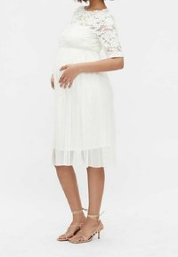 Robe en dentelle blanche avec des manches courtes et une jupe fluide et plissée. Portée avec des sandales à brides beiges. Le mannequin caresse doucement un ventre de femme enceinte.