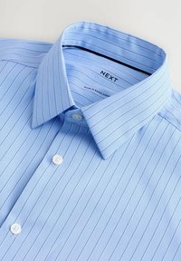 Camicia a righe blu chiaro con colletto classico, caratterizzata da bottoni bianchi e una texture del tessuto liscia. Etichetta "NEXT" visibile all'interno del colletto.