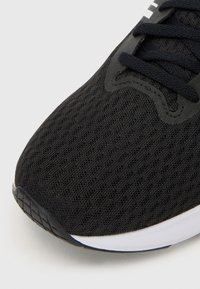 Chaussure de sport noire avec un dessus en maille texturée, bout rond et lacets noirs. La semelle est blanche avec une semelle en caoutchouc noire pour une bonne adhérence.