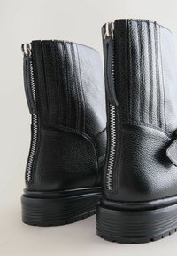 Botas negras hasta el tobillo hechas de cuero texturizado, con cremalleras laterales y detalles acanalados en la parte superior. Suelas de goma negra gruesa para mayor durabilidad.