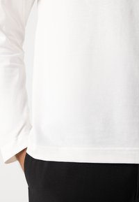 Cămașă albă cu mâneci lungi, cu guler rotund, confecționată dintr-un material de bumbac neted. Marginile se află imediat deasupra pantalonilor negri, evidențiind o siluetă curată.