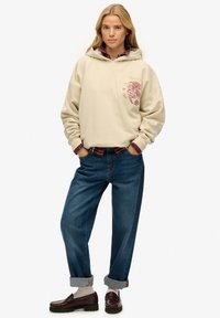 Vrouw staat en draagt een beige hoodie met een rode draakprint, een rood geruit overhemd eronder, opgerolde blauwe spijkerbroek, witte sokken en bruine loafers.