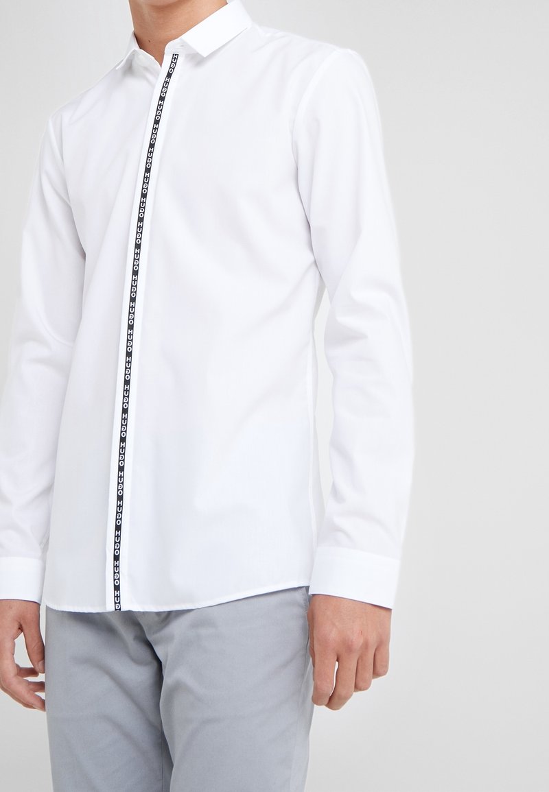 Valkoinen pitkähihaisista paita, jossa on nappikiinnitys edessä ja musta logopainatus placketin reunassa. Sileä kangas ja klassinen kaulus.