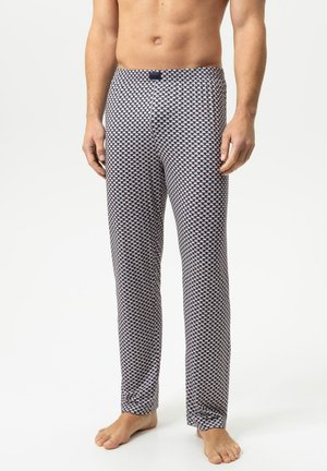 Man die een zwart-wit geruite loungbroek draagt, staat blootsvoets tegen een witte achtergrond.