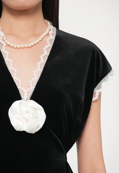 Vestido de veludo preto com decote em V profundo com detalhe em renda, um broche floral de cetim no centro e mangas curtas e esvoaçantes.