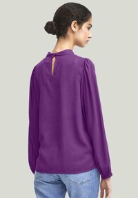 Blusa viola con maniche lunghe, spalle gonfiate, dettaglio a chiave sul retro e texture liscia, abbinata a jeans azzurri chiari.