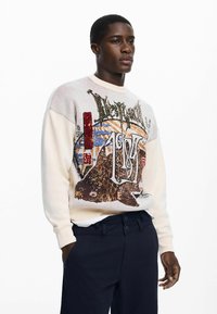 Maglione color crema con tessuto strutturato, caratterizzato da un audace design grafico di un pesce e motivi astratti. Abbinato a pantaloni blu scuro.