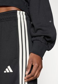 Čierne športové oblečenie je z texturovanej tkaniny s tromi vertikálnymi bielymi pruhmi, logom Adidas na ľavej nohe a detailom rebrovaného lemu.