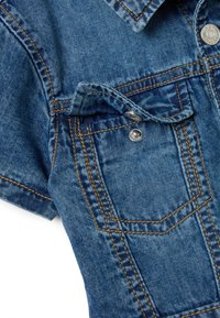 Chemise en denim avec un lavage bleu, des coutures saillantes en orange, une fermeture bouton et une poche poitrine. Tissu texturé avec une finition douce.