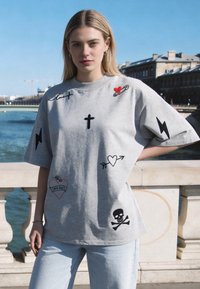 Jeune femme aux cheveux blonds portant un t-shirt gris oversize avec des symboles graphiques noirs et rouges, debout près d'une rivière et d'une rambarde en pierre.