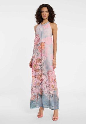 Frau trägt ein ärmelloses, bodenlanges Blumenkleid in Rosa-, Blau- und Grüntönen, kombiniert mit pinkfarbenen offenen Sandalen.