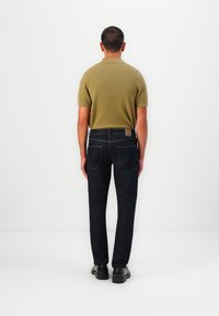 Dunkelblaue, gerade geschnittene Jeans, kombiniert mit einem kurzärmeligen, olivgrünen Poloshirt, von hinten gesehen. Schwarze Schuhe.