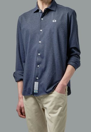 Homme portant une chemise bleu marine à manches longues avec un petit emblème blanc et un pantalon beige, une main dans la poche, sur un fond gris uni.