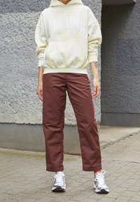 Sudadera cremosa con texto blanco "PEGADOR" y bolsillo canguro. Pantalones rectos marrones y zapatillas grises con detalles naranjas.