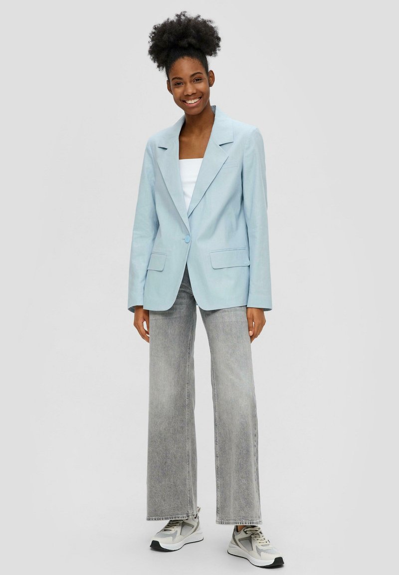 Blazer azzurro chiaro realizzato in un tessuto morbido, con revers a divergenza, chiusura a un bottone e due tasche frontali. Indossato con jeans grigi a gamba larga.