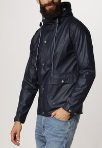 Marinblå vattentät jacka med huva, med silverfärgade tryckknappar, framfickor och dragsnoddar med togglar. Slät, glänsande yta.